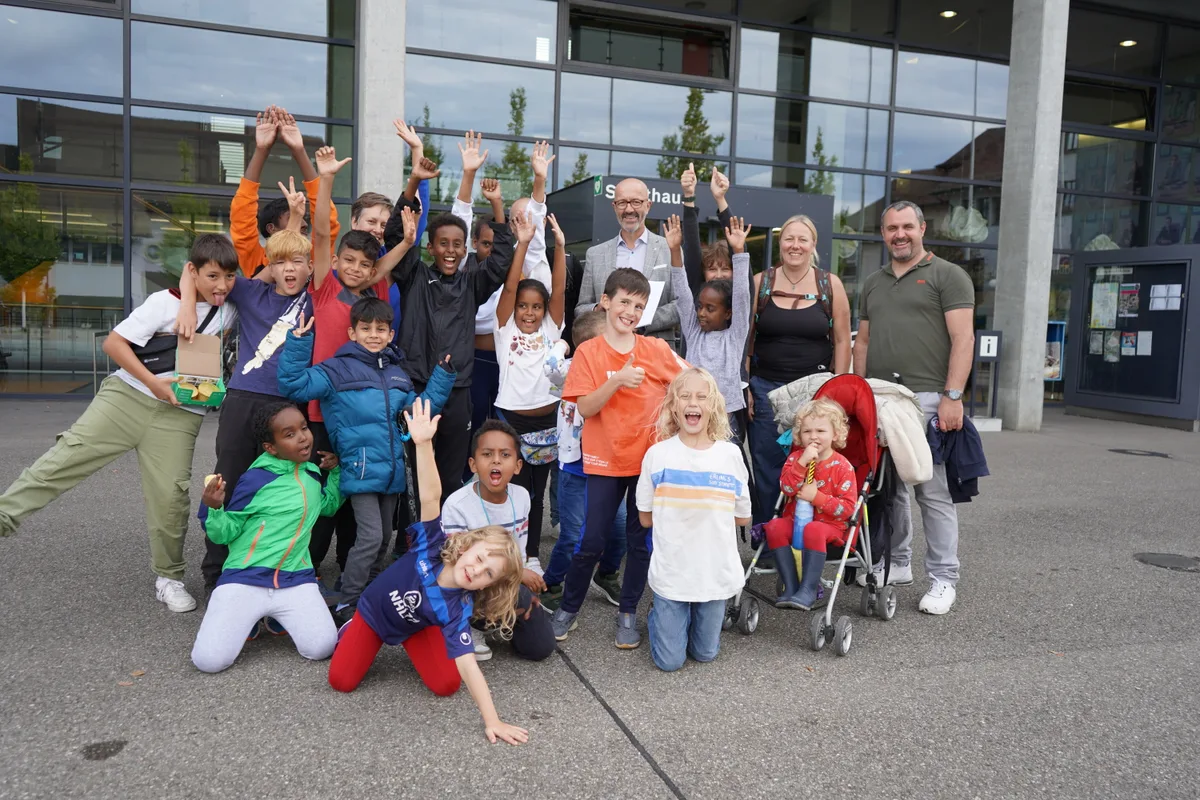 Gruppenbild mit jubelnden Kindern und einigen Erwachsenen vor der Fensterfront eines öffentlichen Gebäudes. 